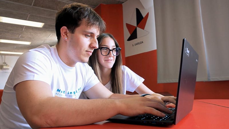 Two people are sitting at a table, working on a laptop. Their faces are blurred for privacy. The person in the foreground is wearing a white t-shirt with the text 'Met ingegneria' and 'SCUOLA TIME TO'. The background features a red wall with a geometric design and the text 'L. GARUDA'.