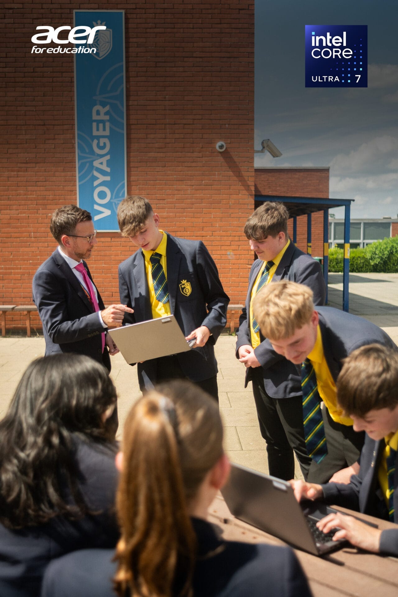 Students in school uniform standing in front of school wall, one of them is holding an Acer TravelMate laptop, and by his side stands a teacher in suite and tie
